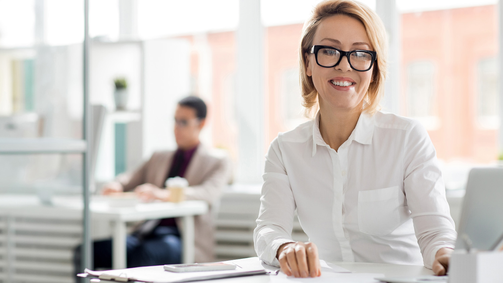 Happy middle aged businesswoman in eyeglasses sitting by her workplace in office and looking at camera with toothy smile